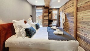 Un dormitorio con techo de madera, una cama con ropa de cama blanca y almohadas decorativas, una mesita de noche con una toalla, y una pared de madera con una bata colgada.