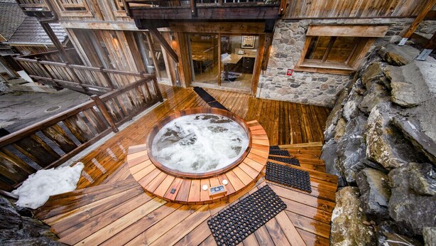 Una bañera de madera rodeada de paredes de piedra y suelo de madera, con un banco de piedra y una escalera de madera cerca.