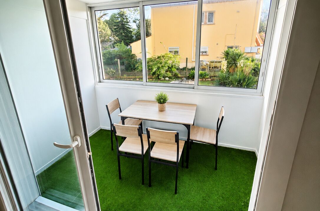 Una habitación con una mesa, sillas, una planta en maceta y vistas a un edificio amarillo a través de ventanas amplias.