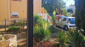 A view through a window of a residential area with a yellow building, parked cars, and a garden with various plants.