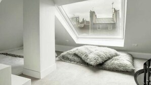 Ein Zimmer mit schrägen Wänden, einem großen Fenster und einem flauschigen weißen Kissen auf dem Boden.