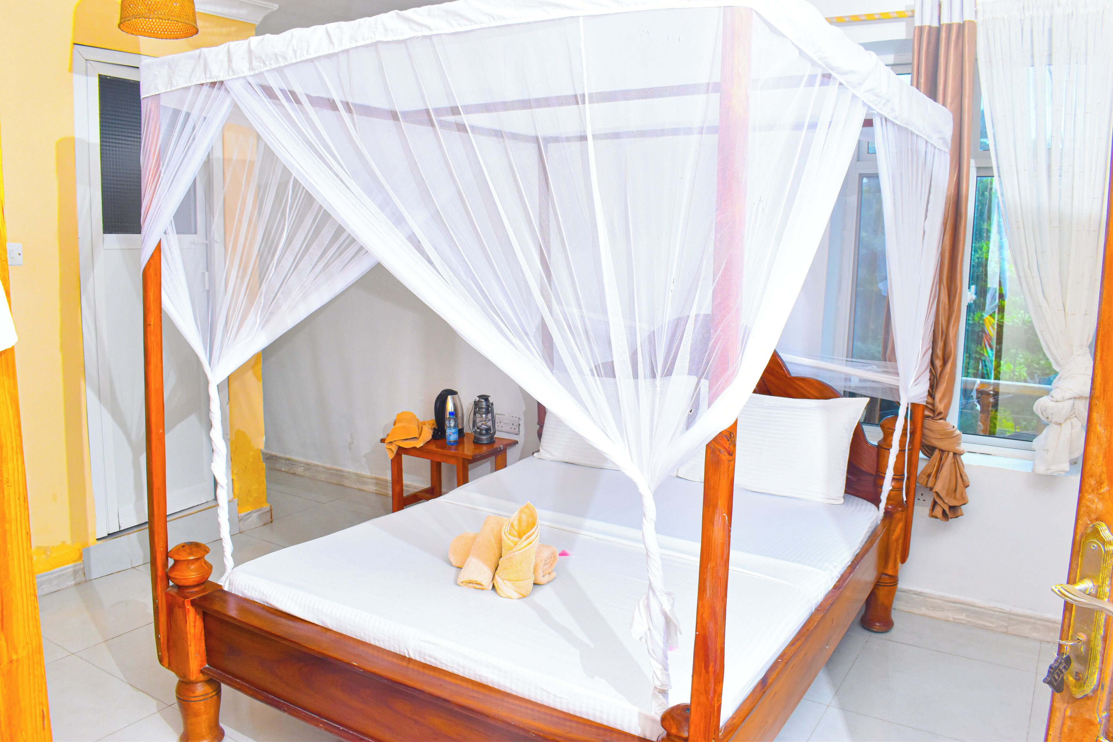 A bed with a canopy, a bedside table with a bottle and a cup, and a window with curtains.