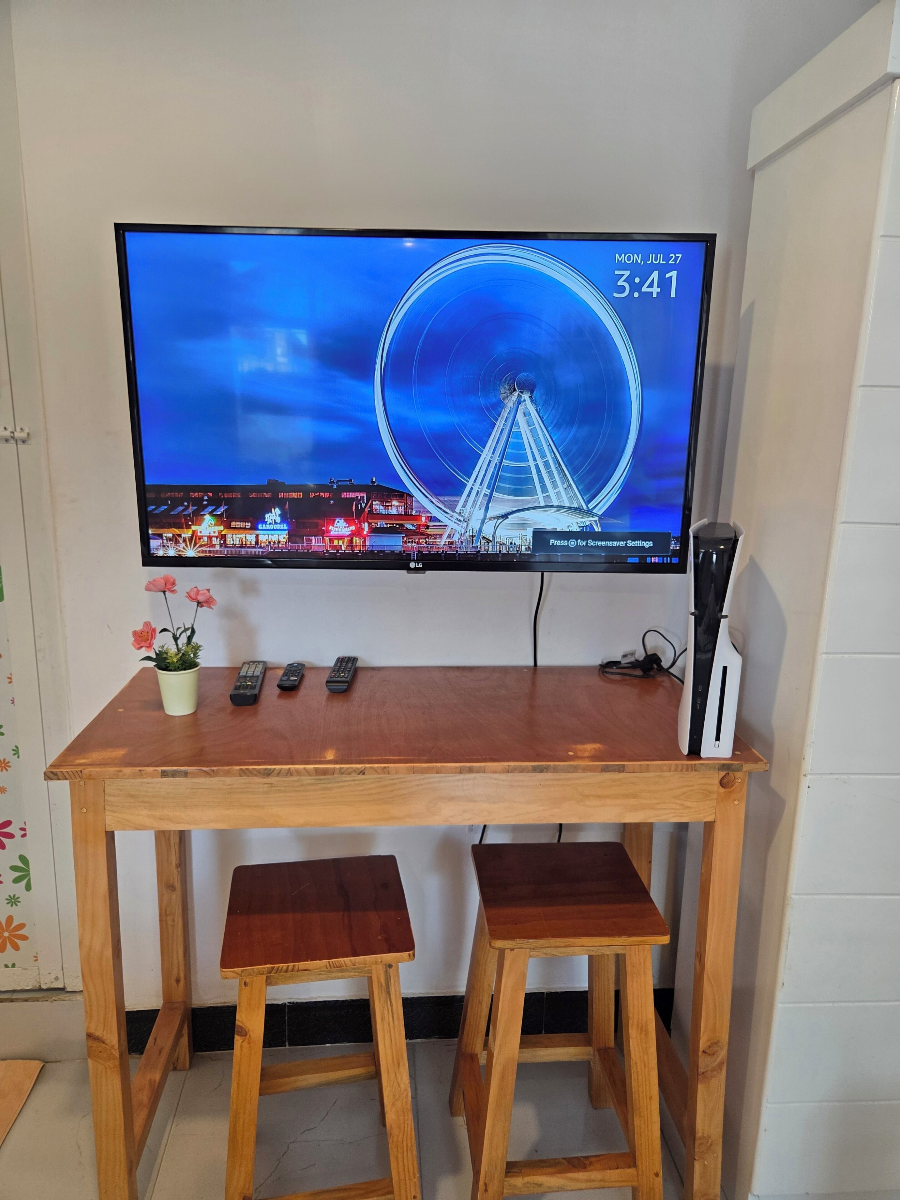 A television mounted on a wooden table with two stools, a remote control, and a gaming console.
