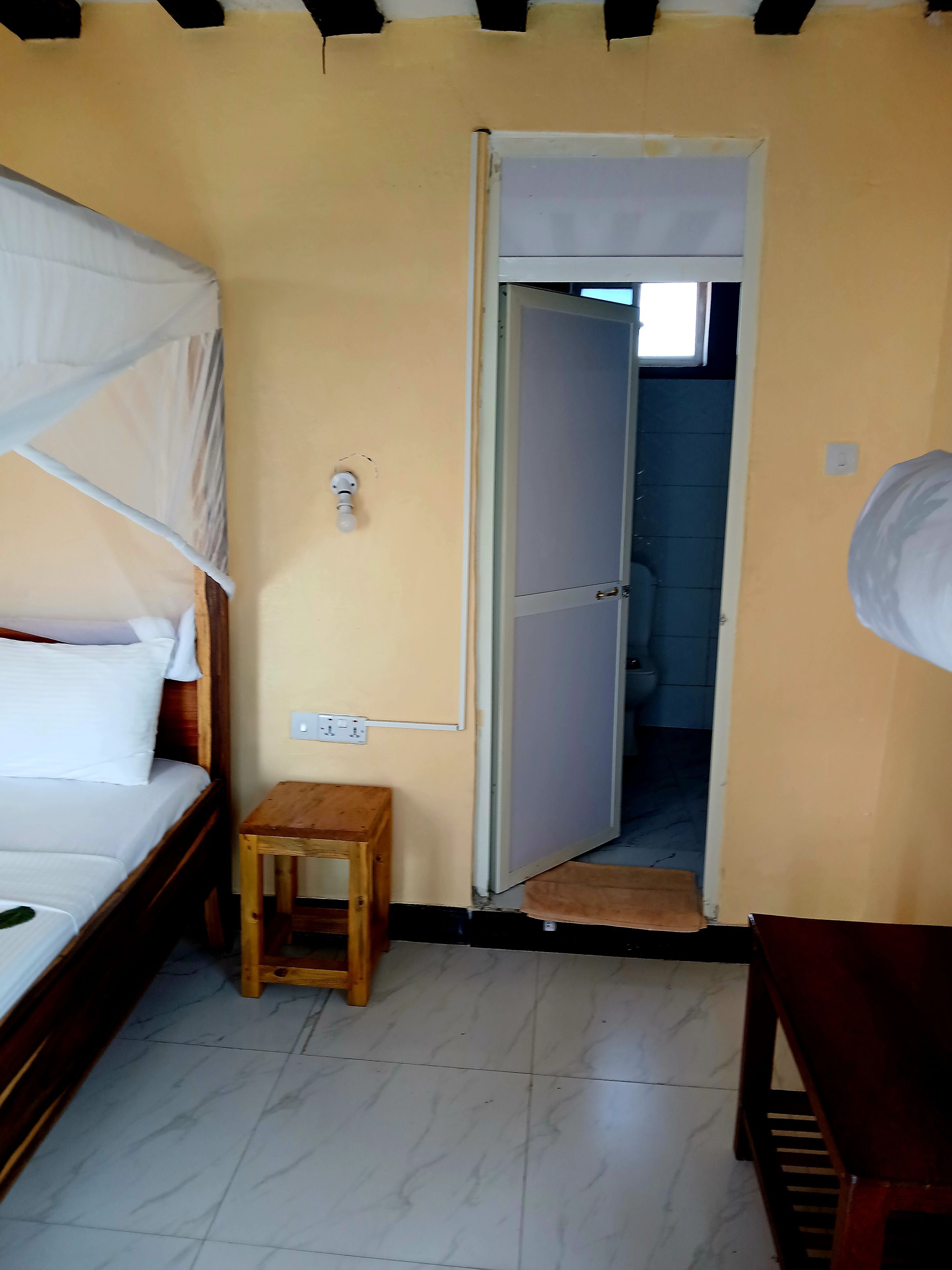 Quarto com uma cama, um banquinho de madeira e uma porta que dá para outro espaço.