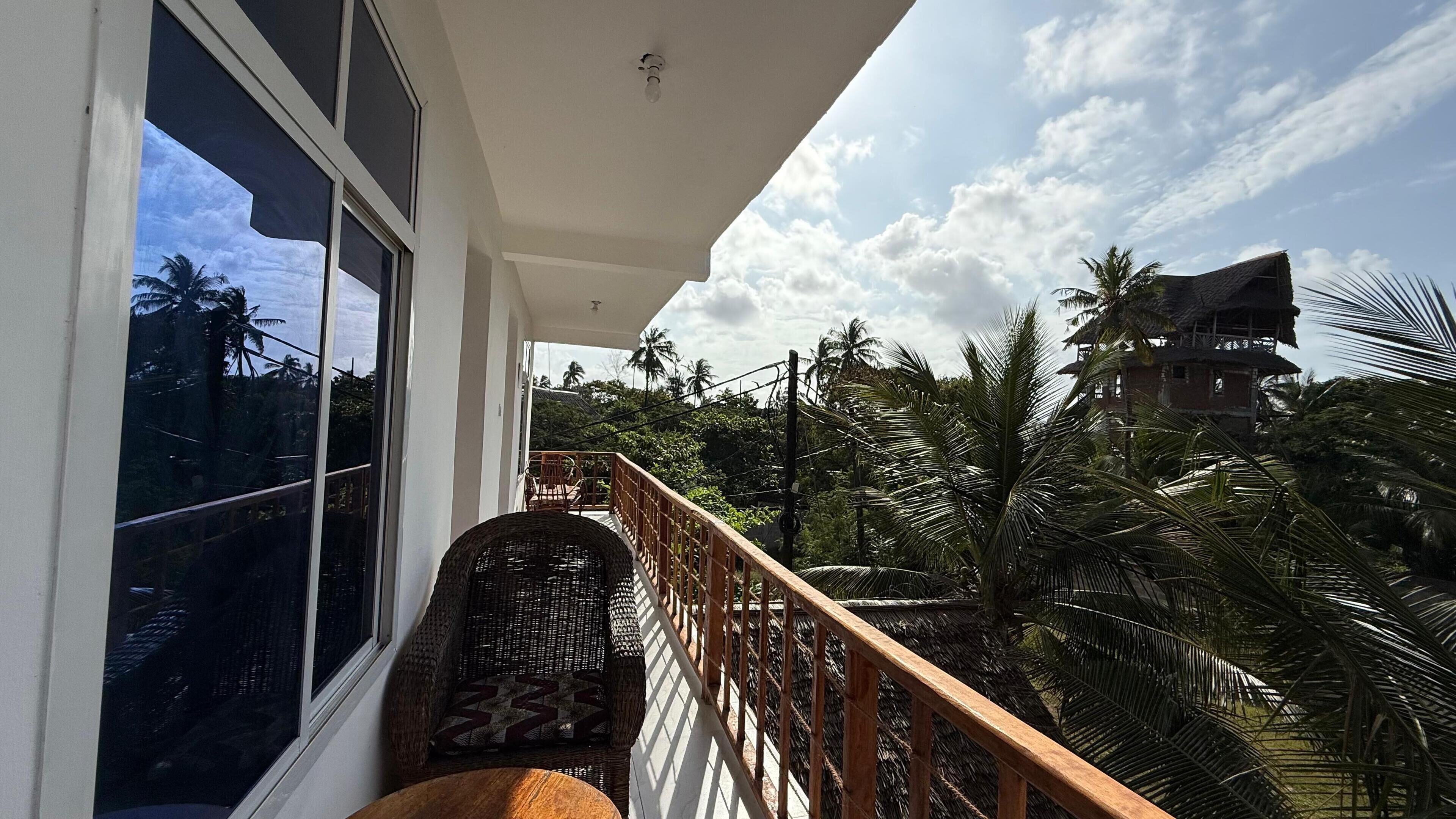 A balcony with a wicker chair and a wooden railing overlooking a tropical landscape with palm trees and a distant building.