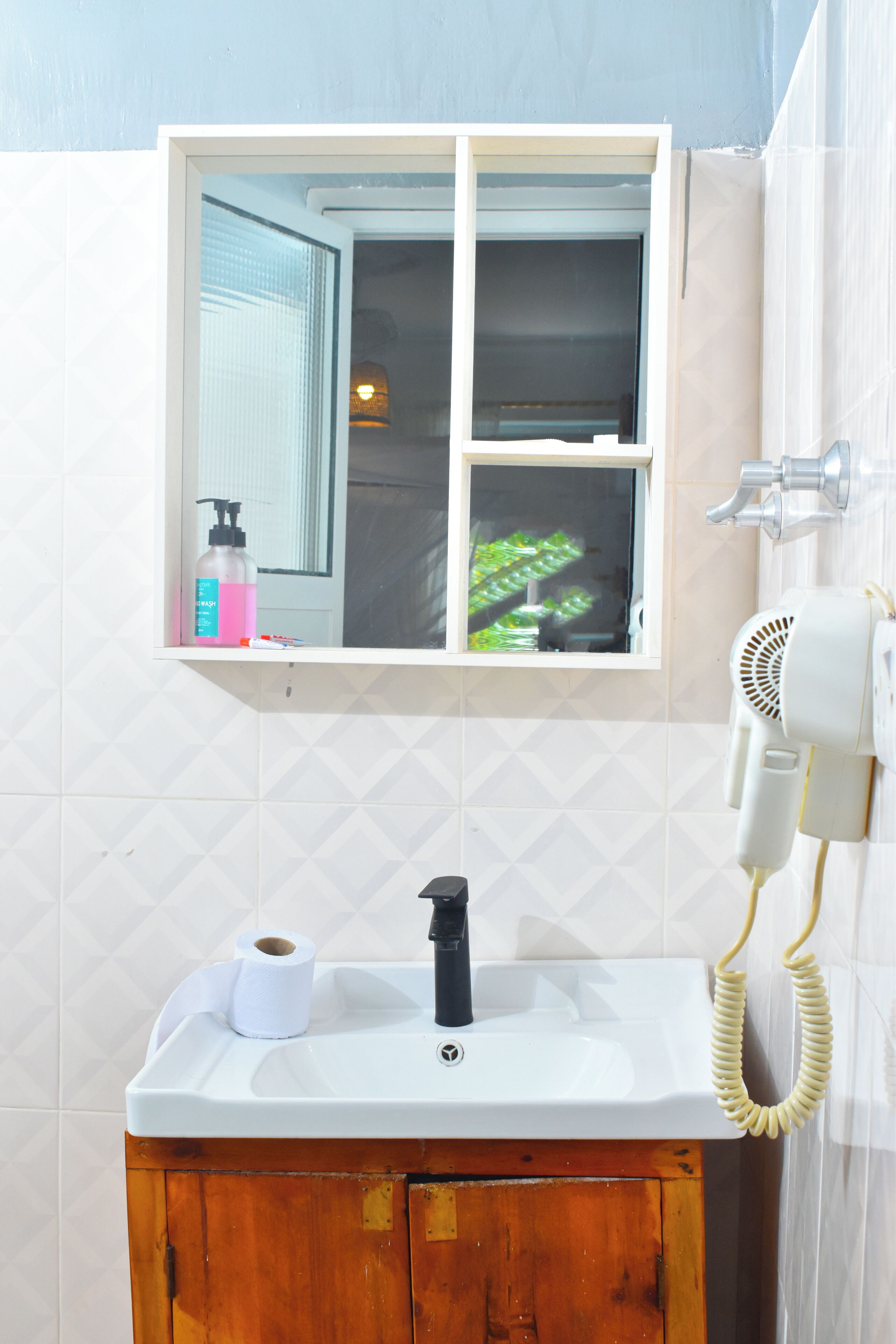 Un baño con ventana, lavamanos, secador de pelo y una botella de champú.