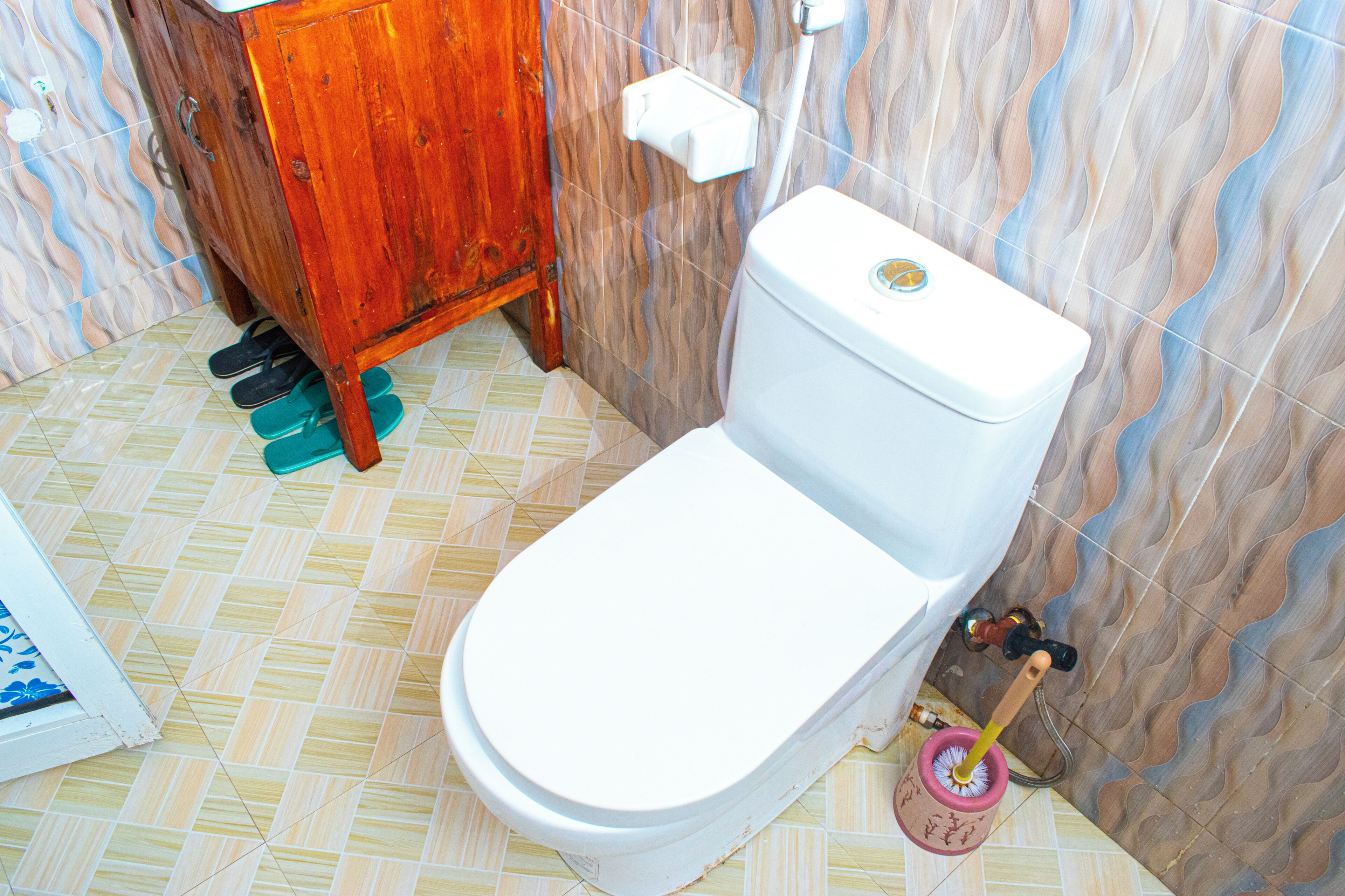 A toilet with a white seat and tank, a wooden cabinet, a mop, and patterned floor tiles.