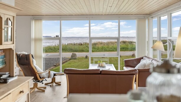Habitación con un amplio ventanal que ofrece vistas al mar y a una carretera. Hay un sofá marr ón, una silla y una mesita de noche con una lámpara.