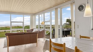 Amplia habitación con techo de madera, ventanas grandes, sofá de cuero marrón, mesa de comedor con sillas y vista al mar.