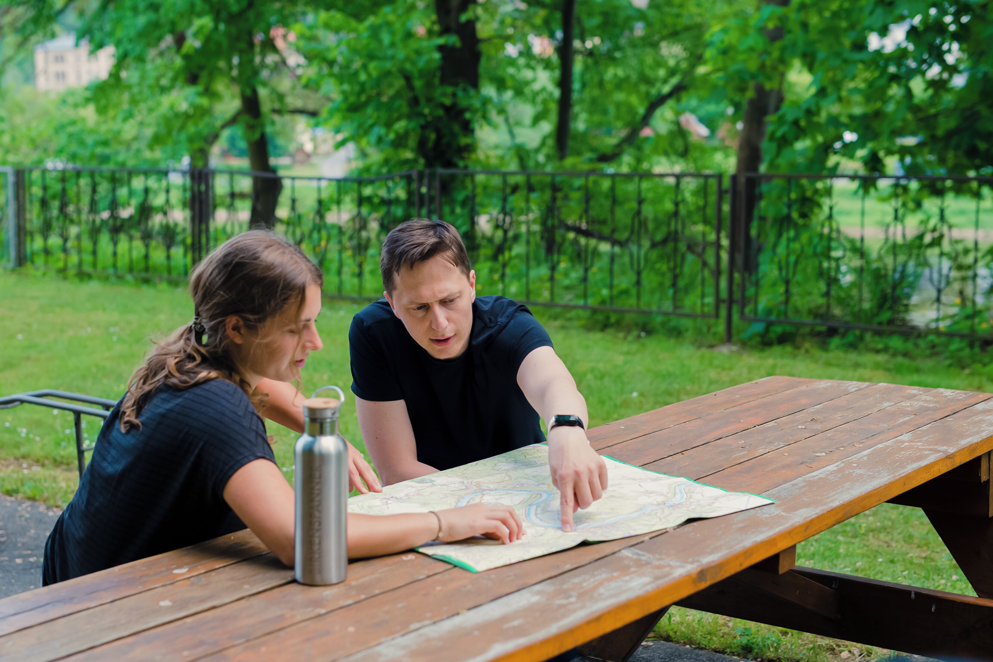 Two people at a wooden picnic table with a map, outdoors with greenery in the background.