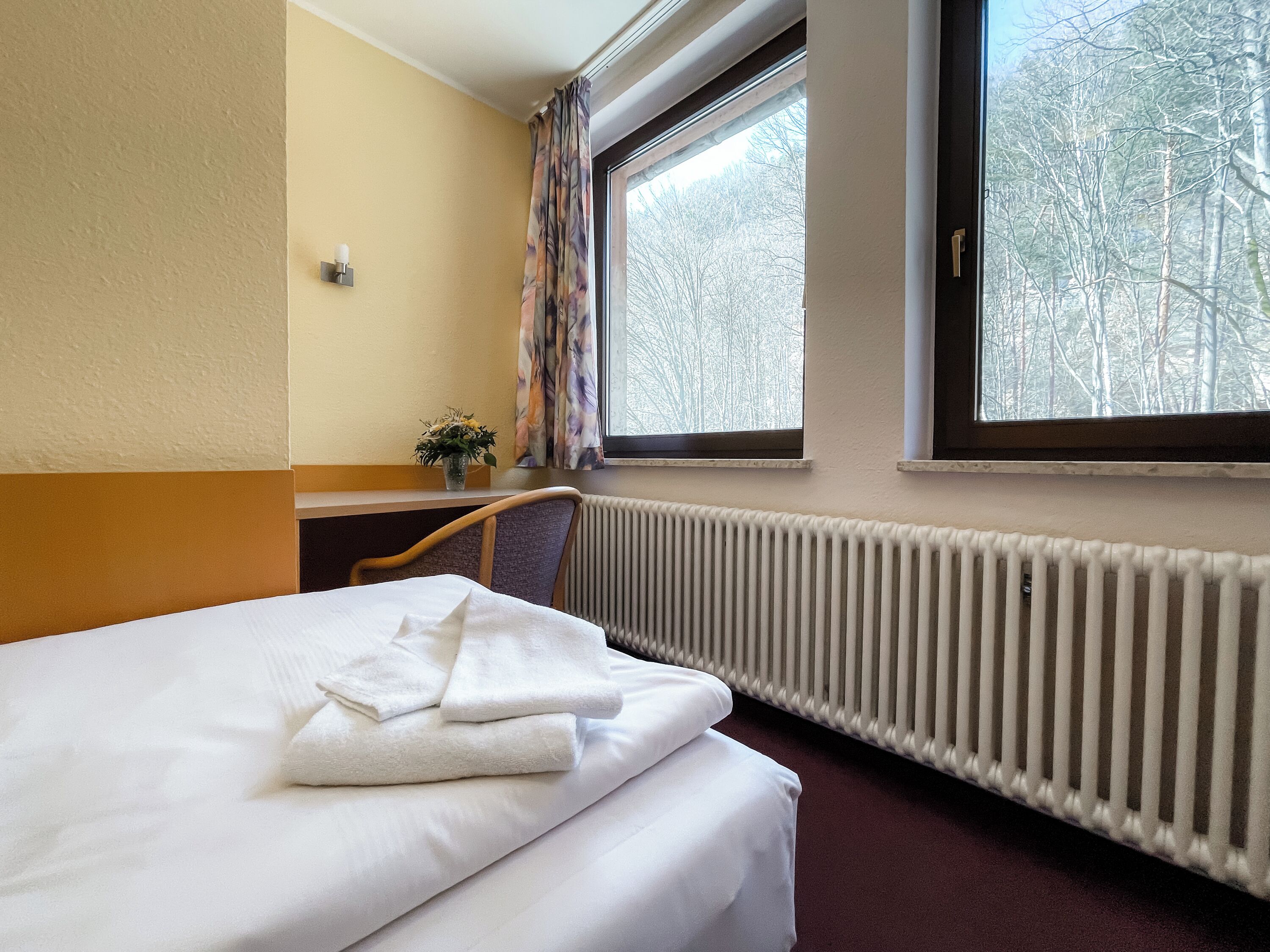 A hotel room with a bed, a window with a view of trees, a radiator, and a small table with a vase of flowers.