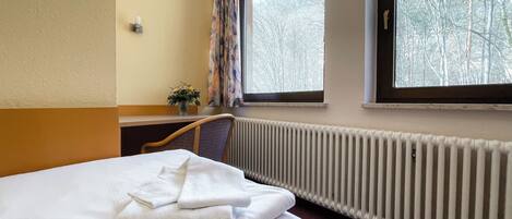 A hotel room with a bed, a window with a view of trees, a radiator, and a small table with a vase of flowers.