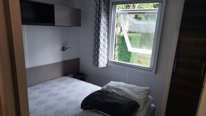A small, single-bed room with a window, a bedside table, and a built-in shelf.