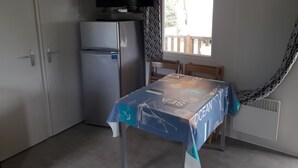 A small room with a table covered in a blue tablecloth with nautical designs, a refrigerator, a television on top of the fridge, and a window with a view of a wooden deck.