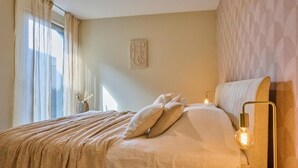 Un lit bien fait, recouvert d’une couverture beige, avec deux oreillers et un luminaire fixé au mur.