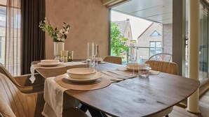 Un coin repas avec une grande table dressée pour quatre personnes, un vase rempli de fleurs et une vue sur l’extérieur grâce à de grandes fenêtres.