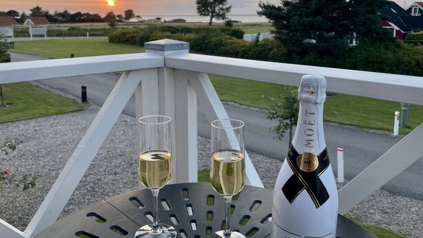 Een balkon met twee glazen witte wijn en een fles Moët champagne, met uitzicht op een zonsondergang boven een kustlandschap.