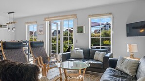 Een ruime woonkamer met een bank, stoelen, een glazen salontafel en een aan de muur bevestigde televisie.
