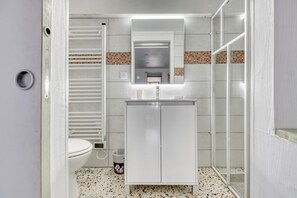 Une salle de bain comprenant des toilettes, un lavabo blanc avec un miroir, un espace douche et un sol carrelé.