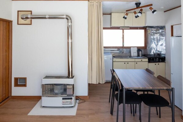 Een kleine keuken met een fornuis, spoelbak en kastjes. Er is een eettafel met stoelen en een kleine verwarming op de vloer.