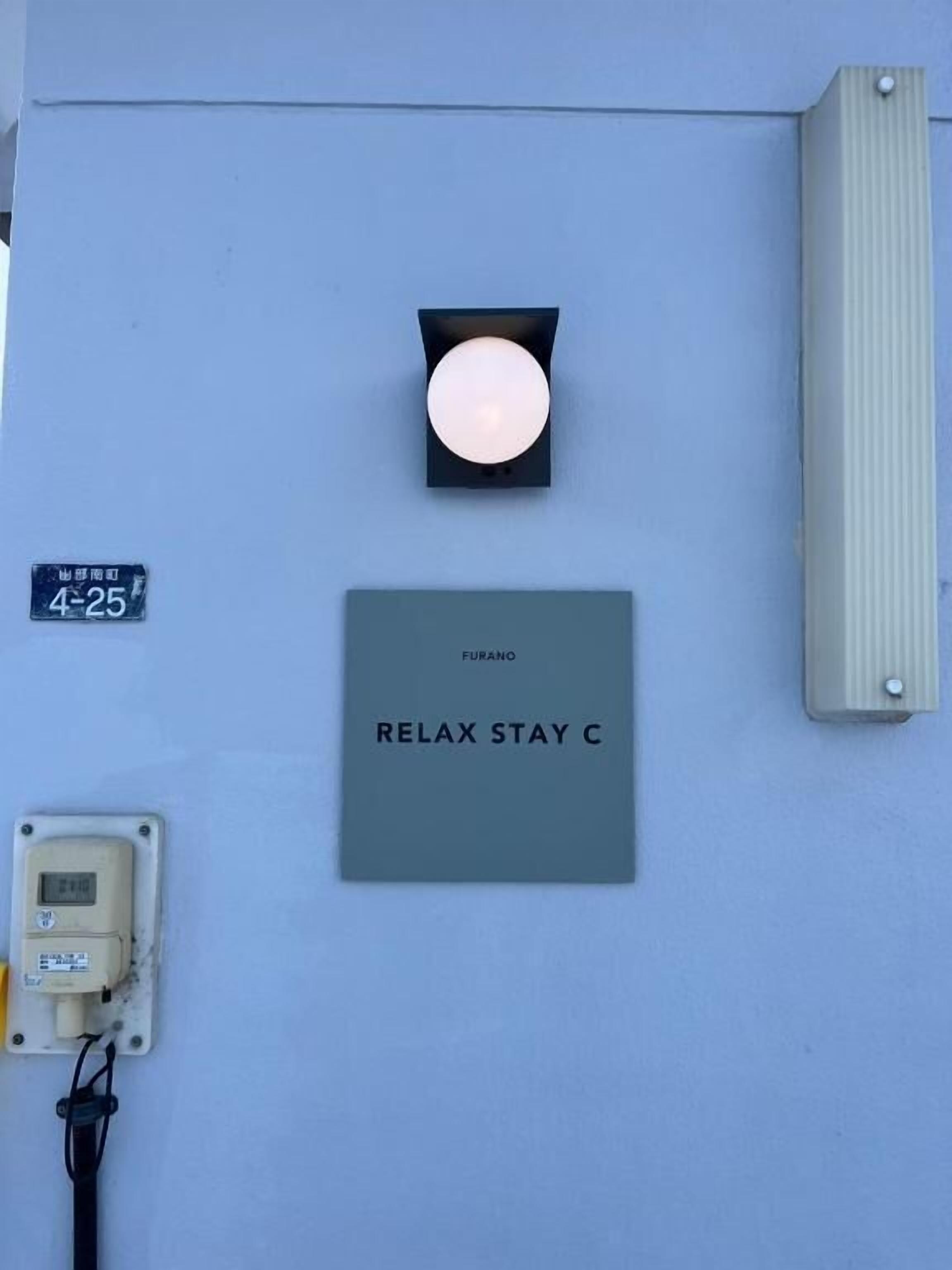 Eine Wand mit einer Leuchte, einem Schild mit der Aufschrift 'FURANO RELAX STAY C' und einem Verteilerkasten.