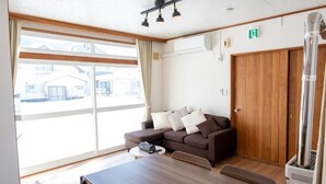A room with a large sliding glass door, a brown sofa with cushions, a wooden table, and a heater. - FURANO RELAX STAY BC (Furano)
