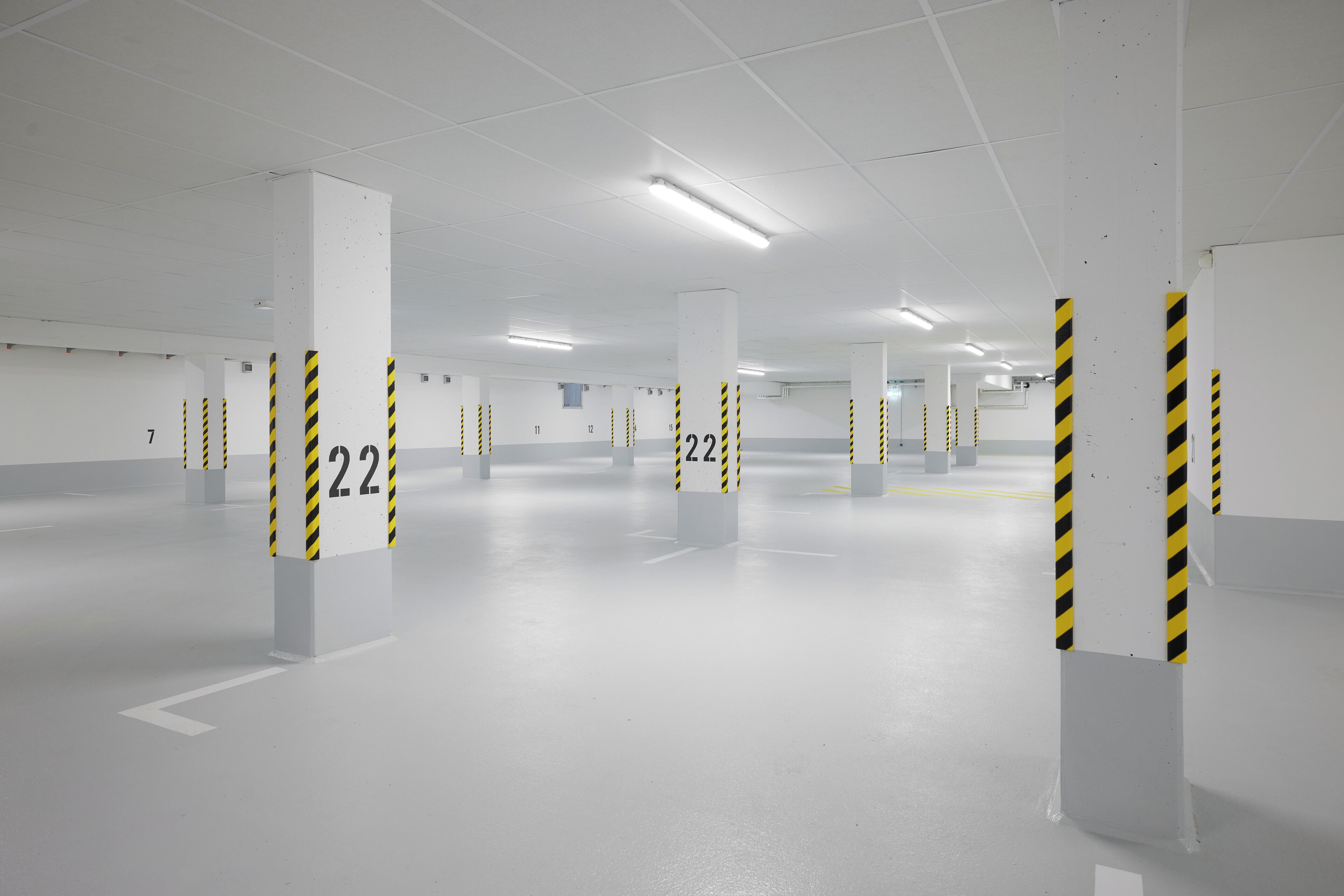 A clean, empty parking garage with white walls and floors, marked parking spaces, and safety bollards with yellow and black stripes.