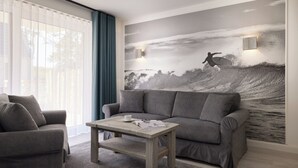 A living room with a grey sofa, a wooden coffee table, and a large wall mural of a surfer. - Completion 2025: Fantastically beautiful apartment for up to 2 people! (Sankt Peter-Ording)