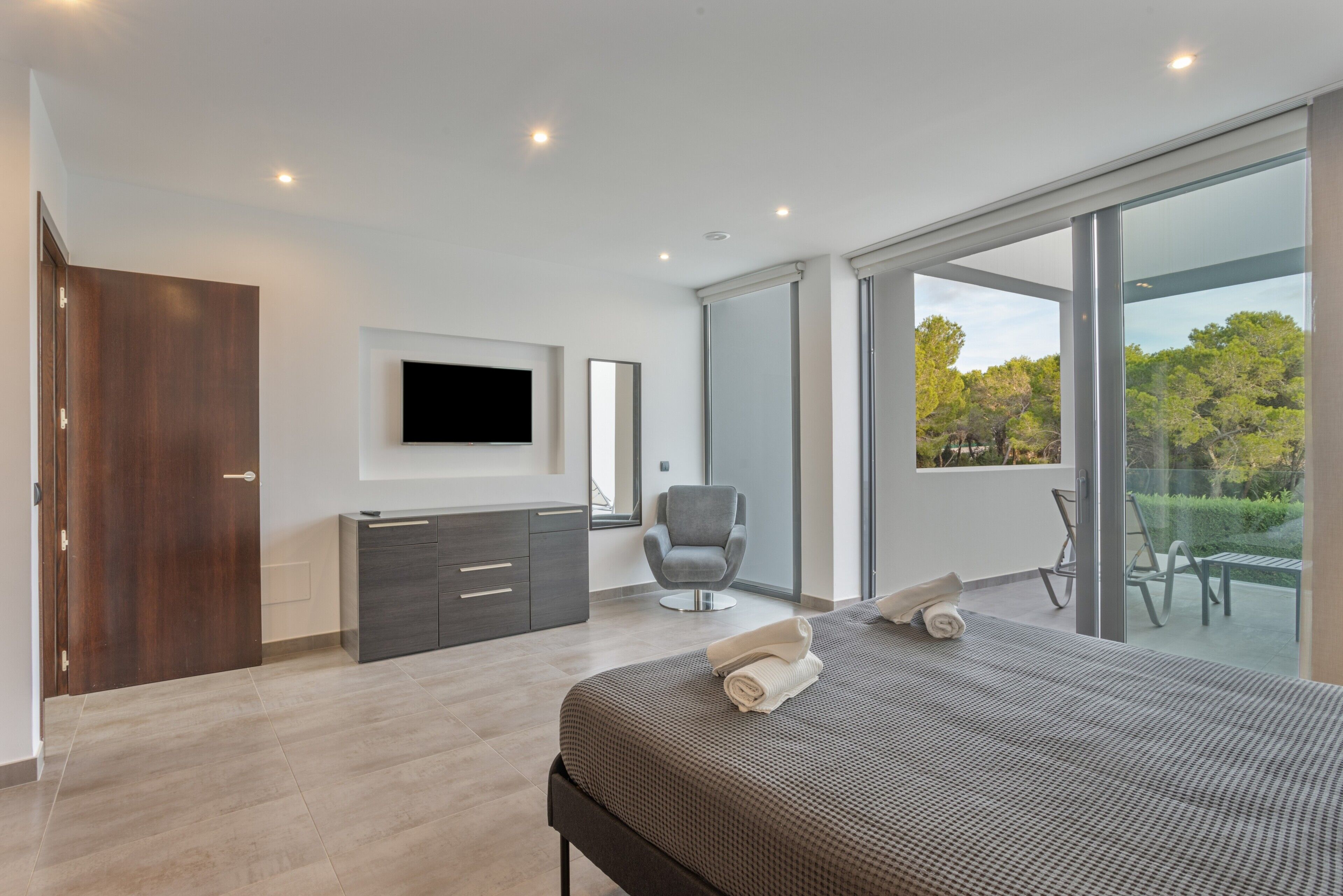 Un dormitorio moderno con una cama, un televisor, una silla y un ventanal corredera a un balcón.