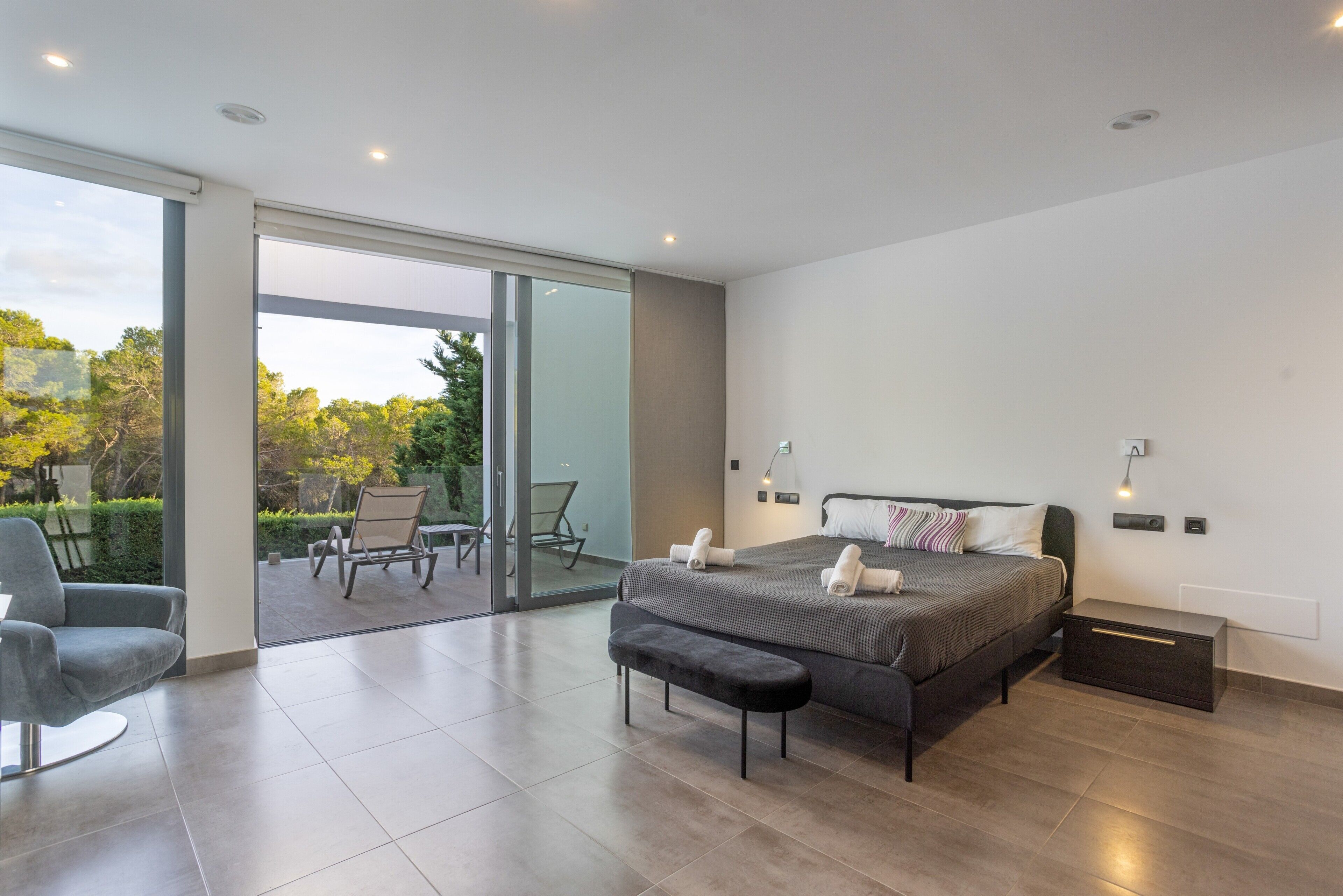 A modern bedroom with a large bed, a bench, a chair, and a sliding glass door leading to an outdoor area.