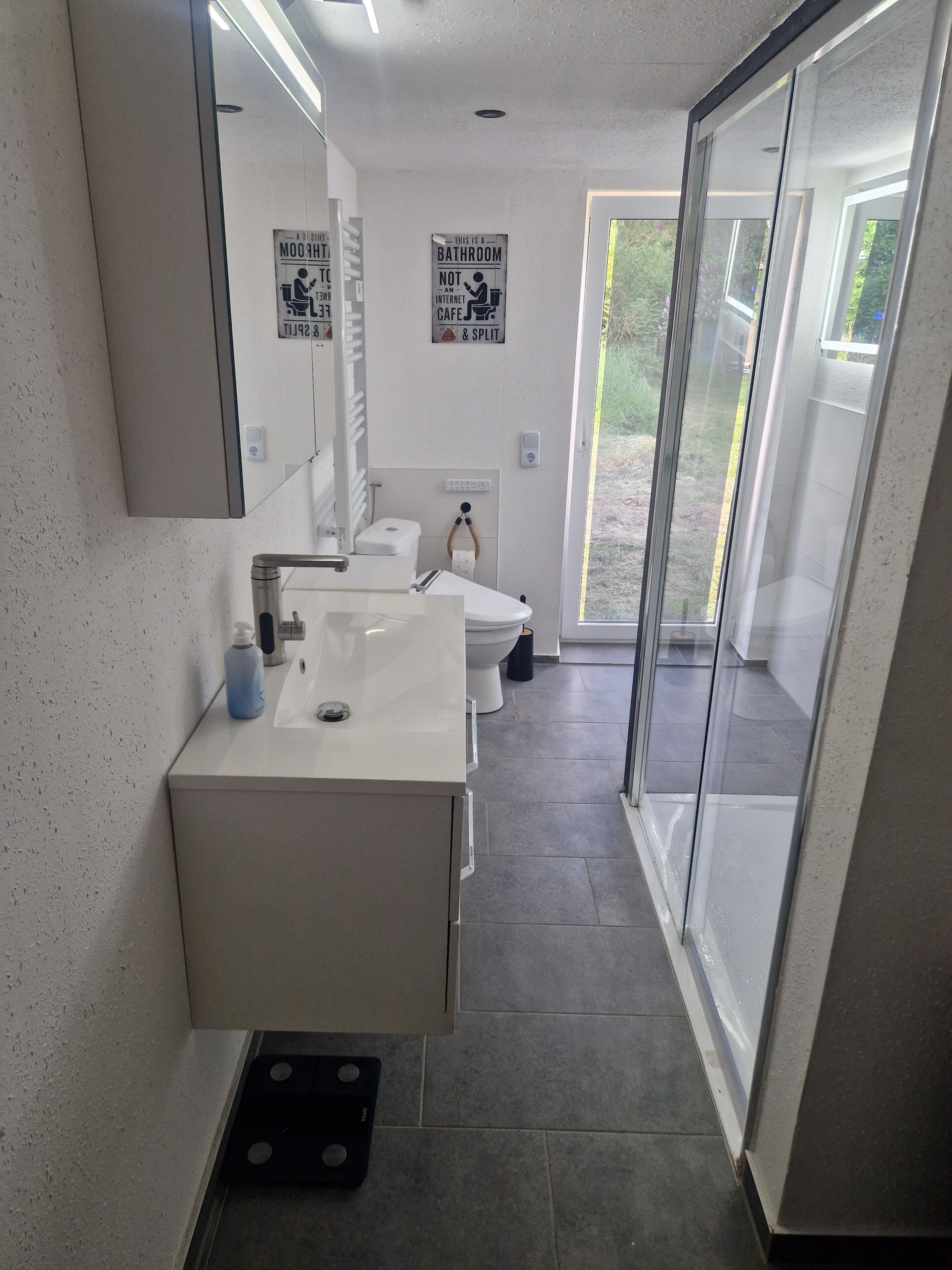 Ett modernt badrum med ett vitt handfat, toalett och duschutrymme. Det finns två skyltar på väggen, en som visar att det är ett badrum och den andra som anger att det inte är ett internetcafé.