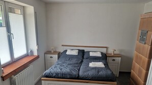 A bedroom with a bed, two bedside tables, a window with blinds, a radiator, and a wooden cabinet.