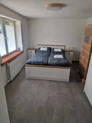 Ein Schlafzimmer mit einem Bett, zwei Nachttischen, einem Fenster mit Jalousien, einem Heizkörper und einem Holzschrank.