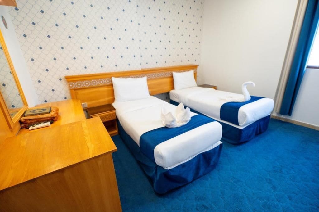 Ein Hotelzimmer mit zwei Betten, jeweils mit blauer Bettdecke und weißen Kissen, einem Holz-Kopfende und einem kleinen Holztisch mit einem Buch und einer Fernbedienung.