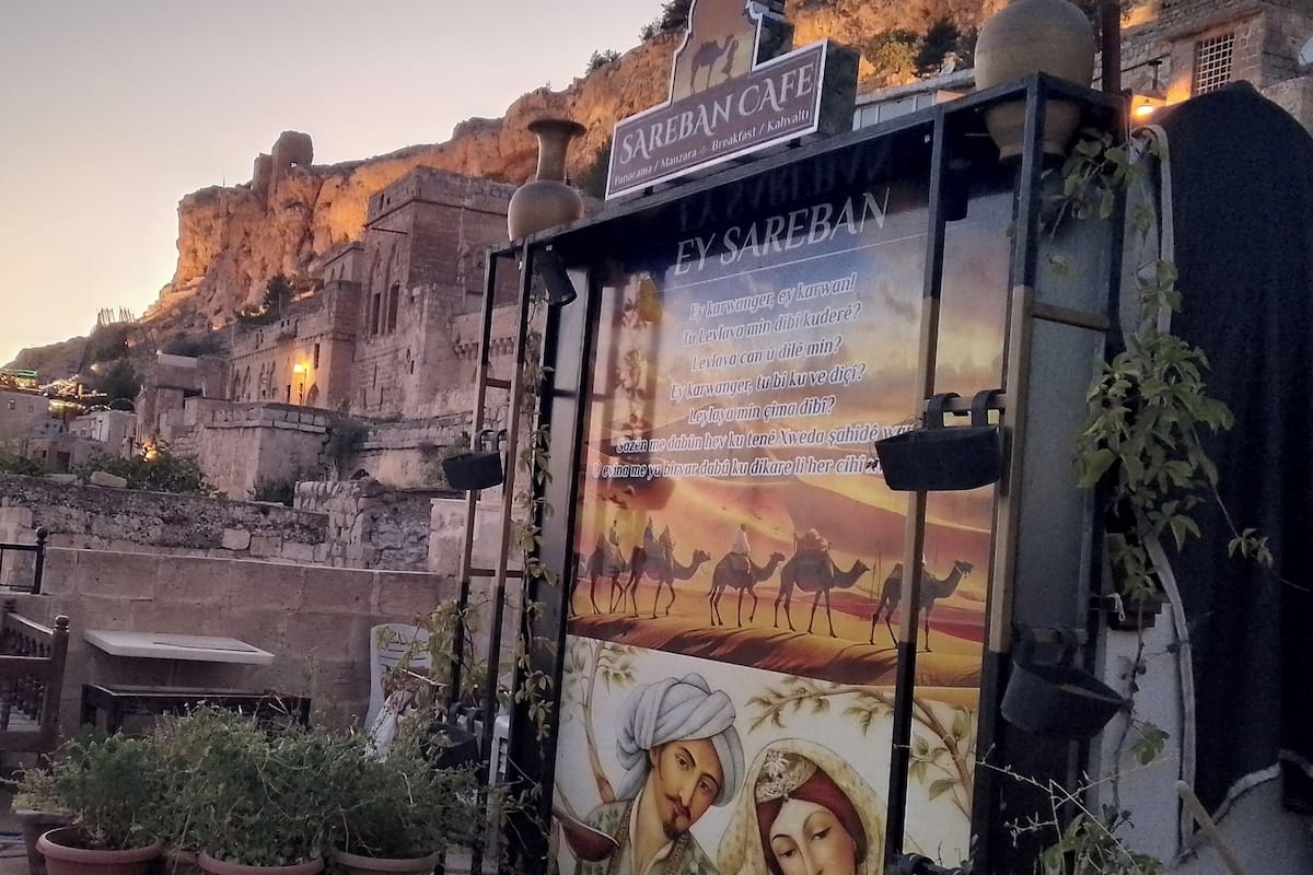 Egy hagyományos épület, amelyen egy „SAREBAN CAFE” feliratú tábla és egy sivatagi tájat ábrázoló falfestmény látható.