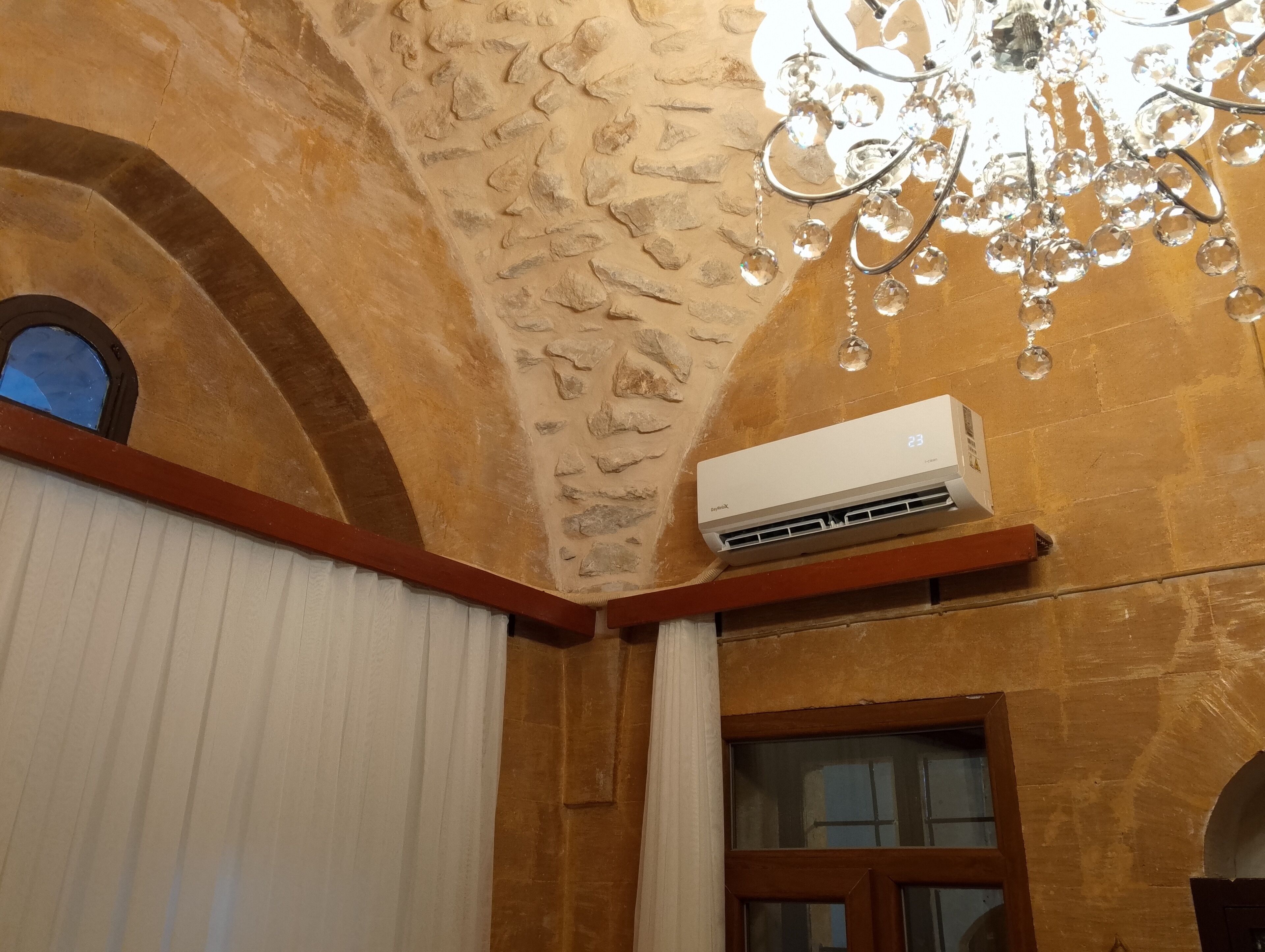 Une pièce avec un mur en pierre, un climatiseur fixé au mur, une fenêtre avec des volets en bois et un lustre.