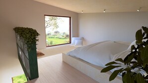 A modern bedroom with a large bed, a view of a garden through a window, and a potted plant.
