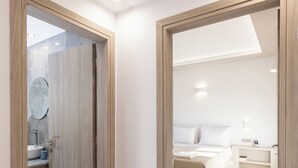 Une chambre d’hôtel moderne avec une porte en bois, une salle de bain dotée d’un miroir rond, un lit avec du linge de lit blanc et une table de chevet.