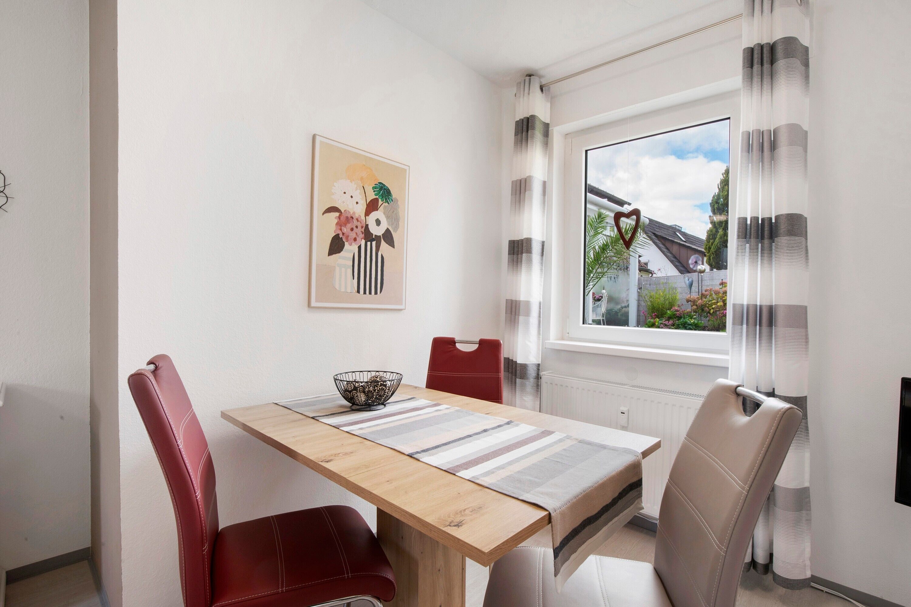 A dining area with a wooden table, red and beige chairs, a striped table runner, a bowl on the table, and a window with curtains.