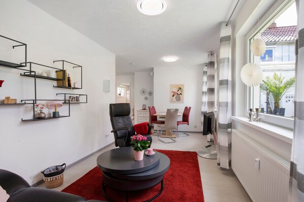 A modern living room with a black leather sofa, a round coffee table with a red rug, and a dining area with a red chair.