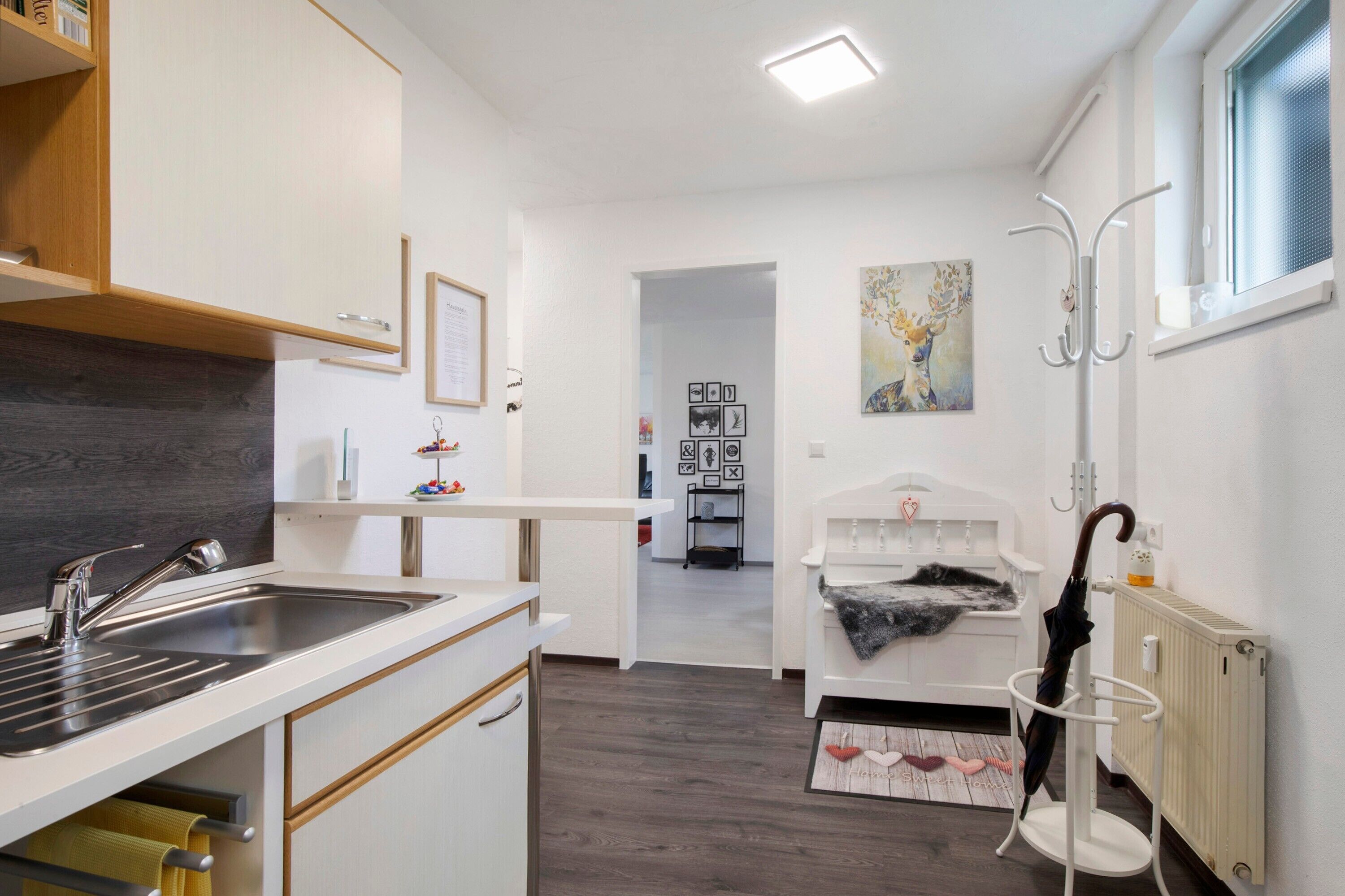 Cocina pequeña con lavamanos, encimera blanca y armario de madera. Un pasillo conduce a un dormitorio con cama y radiador.
