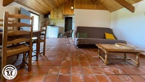 Een rustiek interieur met houten plafond, betegelde vloer, een bank, een eettafel en een keukenhoek.