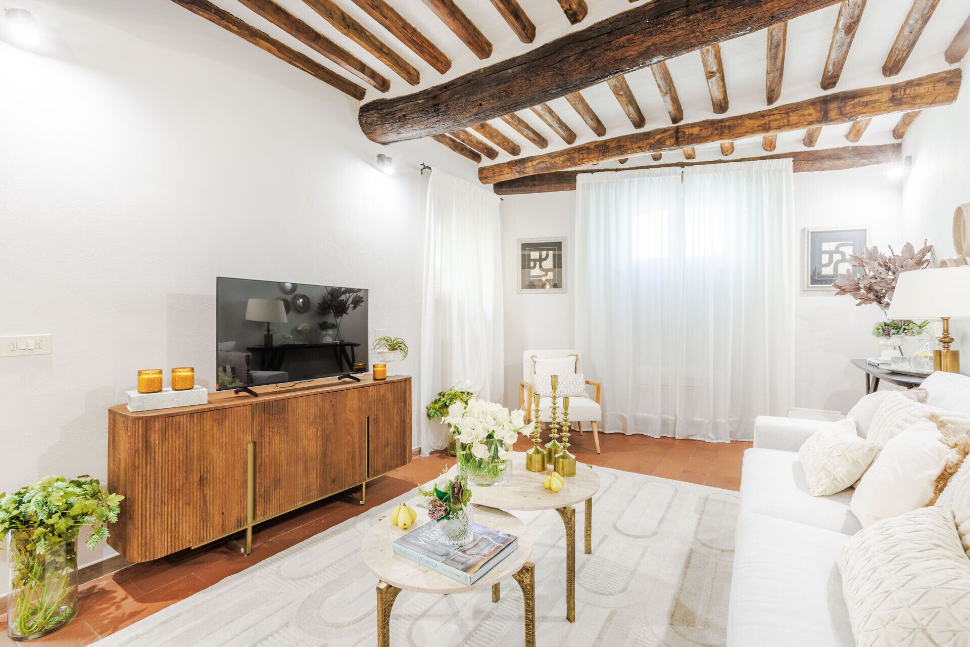 A modern living room with a wooden ceiling, a TV on a wooden cabinet, a white sofa, and a coffee table with books and flowers.