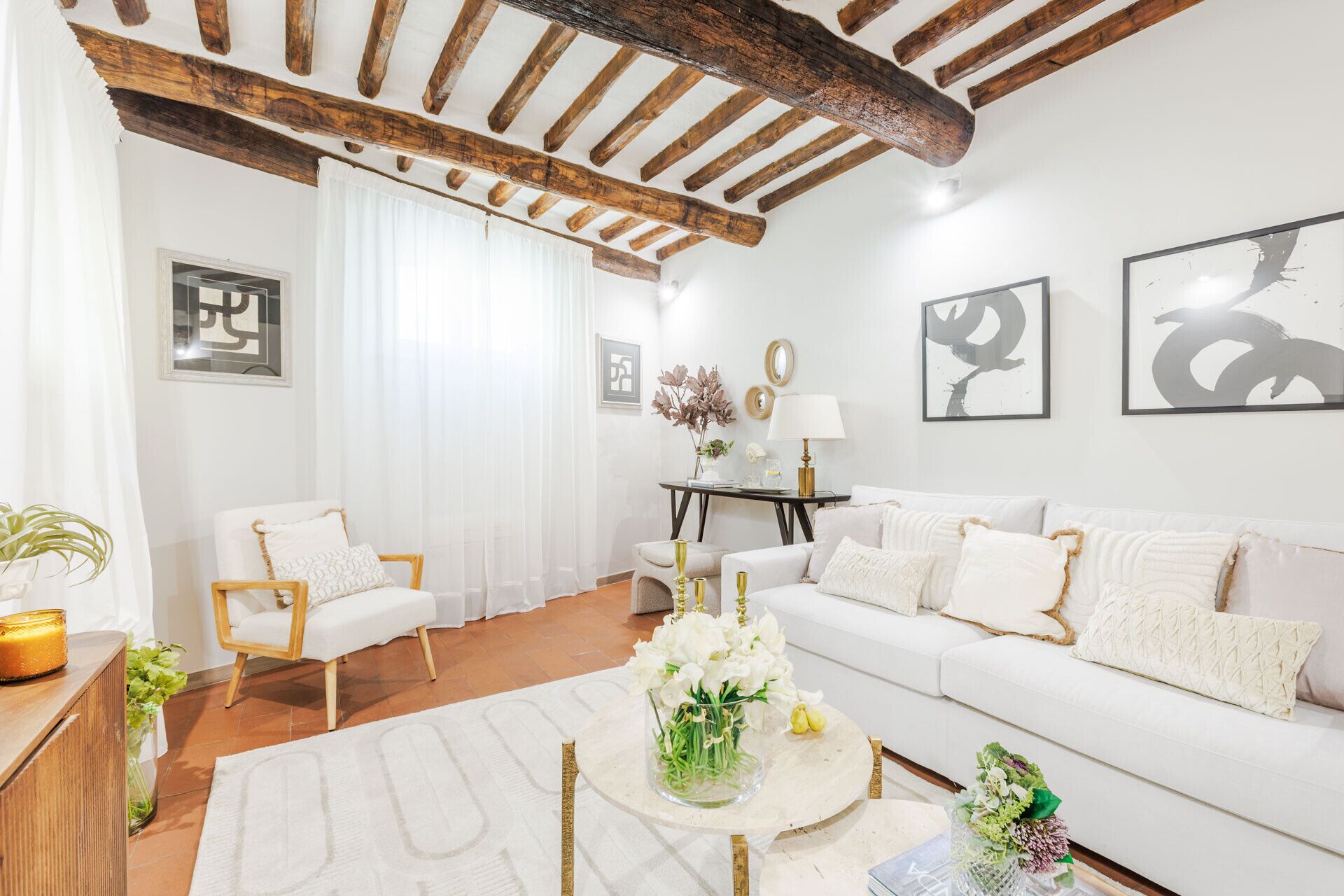 Un salon avec un canapé blanc, une chaise en bois, une table basse ornée de fleurs et un plafond avec des poutres apparentes.