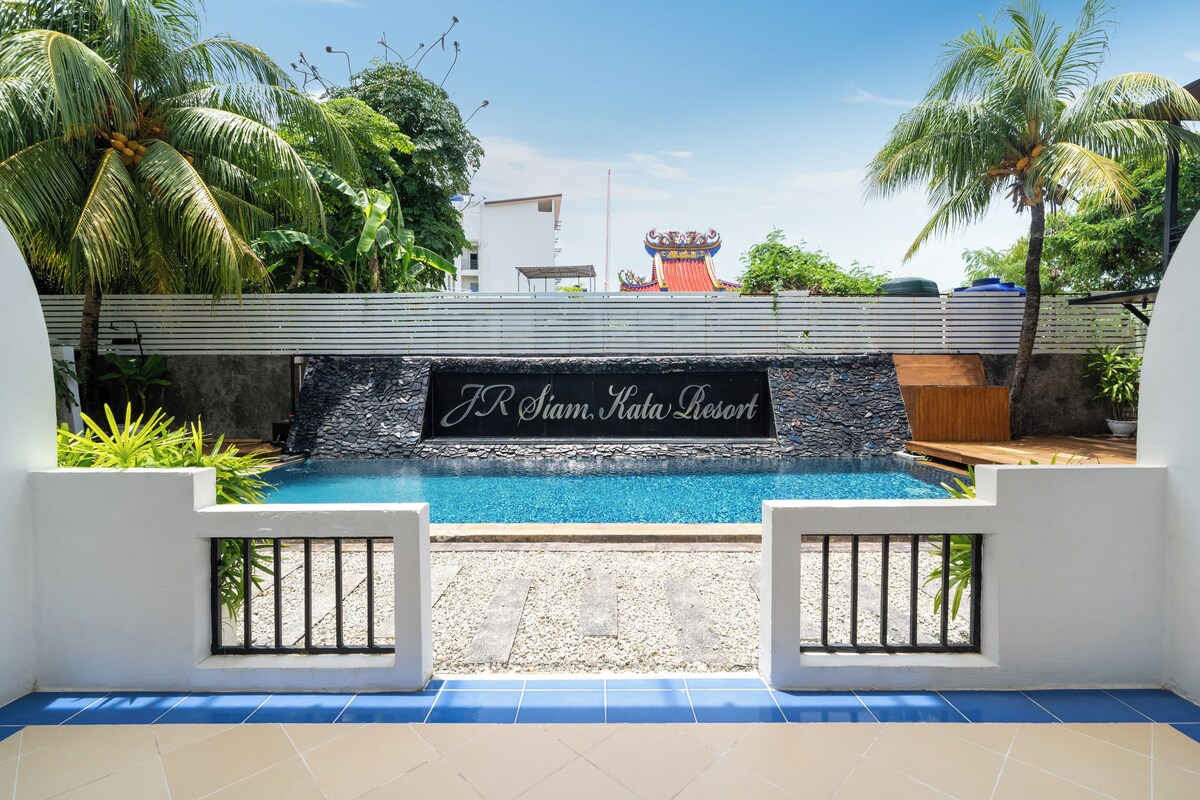 A swimming pool with a sign reading "J R Siam, Kata Resort" surrounded by palm trees and a white building in the background.