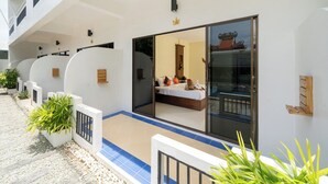 A hotel with a modern design, featuring a large glass door leading to a bedroom with a bed, pillows, and a nightstand. There are potted plants along the walkway and a tiled floor. - JR Siam Hotel (Karon)