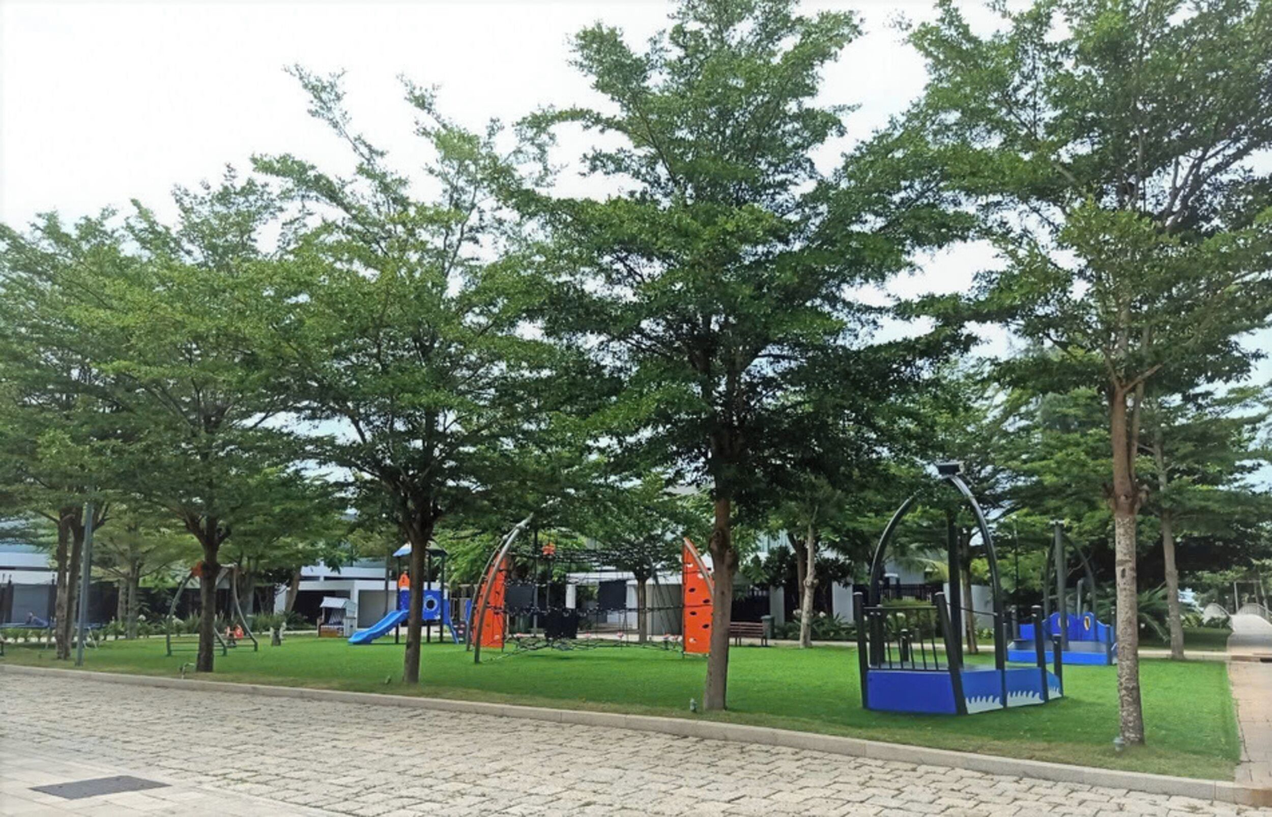 A playground with various play structures, surrounded by trees and a paved area.