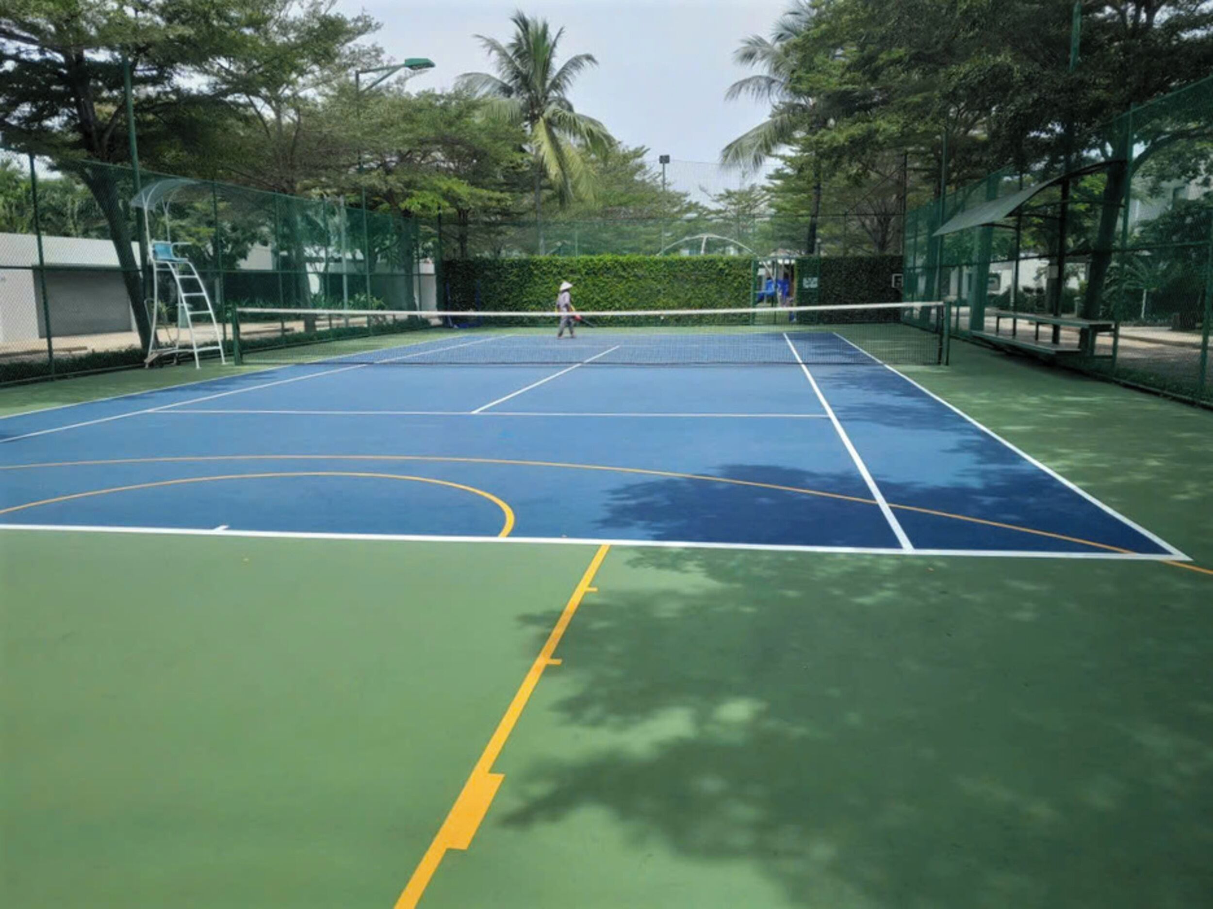 Un court de tennis en plein air, doté d’un filet, entouré d’une clôture verte et d’arbres.