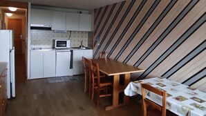 A compact kitchen with white cabinets, a microwave, and a small refrigerator. There is a wooden dining table with chairs and a patterned tablecloth. The room has a striped wallpaper and a wooden floor. - Spacious Apartment in Super Besse for 6 with WIFI (Besse-et-Saint-Anastaise-Super Besse)