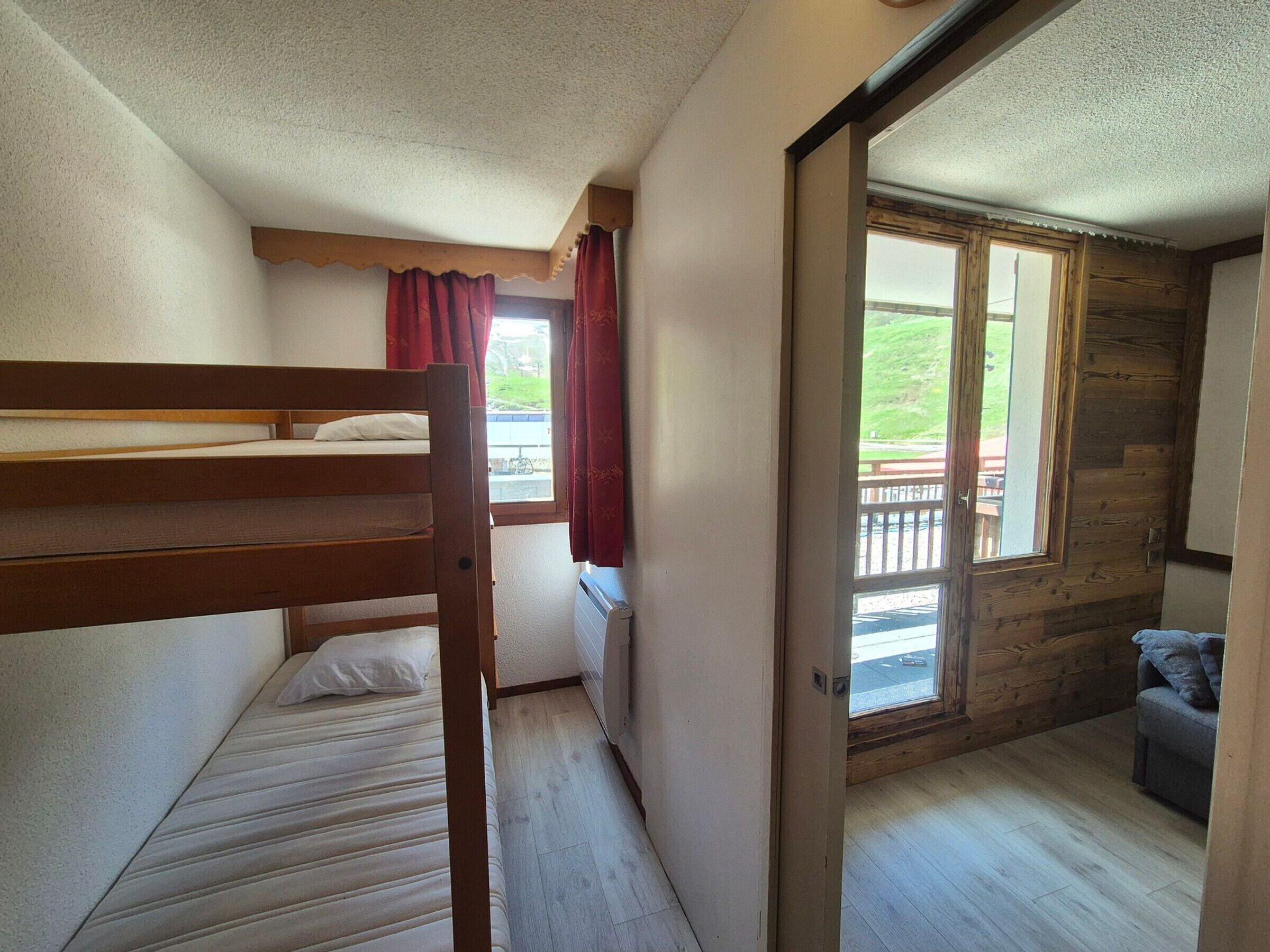 A small, cozy room with a bunk bed, a single bed, a window with curtains, and a view of a balcony.
